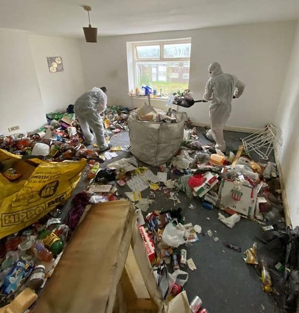 Two workers in protective gear clean up a rubbish strewn room